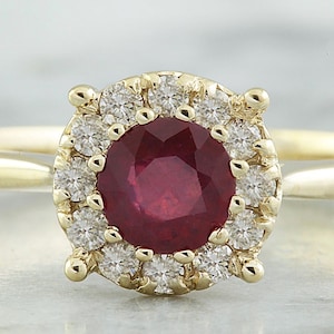 May include: A gold ring with a central, round, deep red ruby, encircled by a halo of small diamonds. The ring's band is plain gold. The gemstones are set in a square setting. The ring is a classic design.