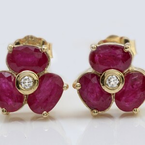 May include: A pair of gold earrings with a flower design. Each earring features three ruby stones and a single diamond in the center.