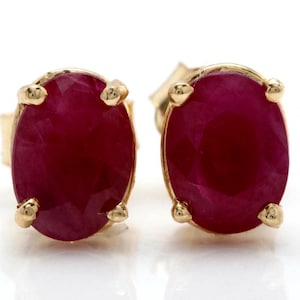 May include: A pair of ruby stud earrings. Each earring features a vibrant, oval-cut ruby gemstone set in a gold-colored metal. The gold setting has four prongs securing the ruby. The earrings are displayed against a white background.