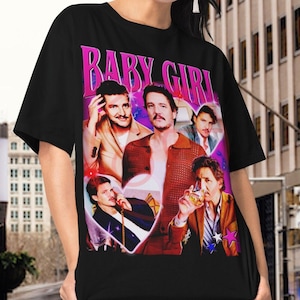 Peut inclure: T-shirt unisexe noir avec un imprimé graphique d'un groupe de personnes en forme de cœur. Le texte "BABY GIRL" est imprimé en lettres roses sur le t-shirt.