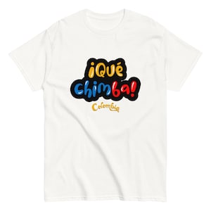 Op de afbeelding: Wit T-shirt met de kleurrijke tekst "¡Qué chimba! Colombia". De tekst is geel, blauw en rood met een zwarte omlijning. Het woord "Colombia" staat in gouden letters onder de hoofdtekst.