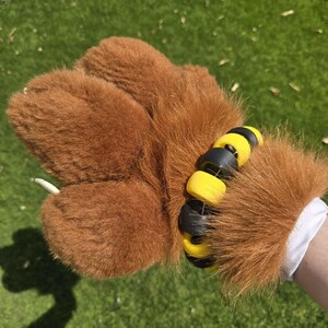 May include: Brown furry hand with black and yellow wrist cuffs. The cuffs are made of multiple round pieces connected together.