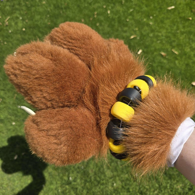 May include: Brown furry hand with black and yellow wrist cuffs. The cuffs are made of multiple round pieces connected together.