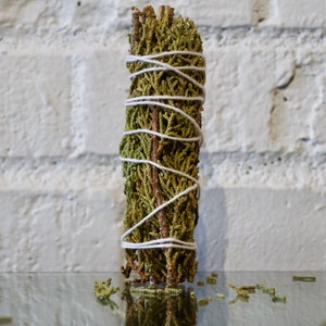 May include: A bundle of dried sage tied with white twine. The sage is green and brown and is arranged in a cylindrical shape.