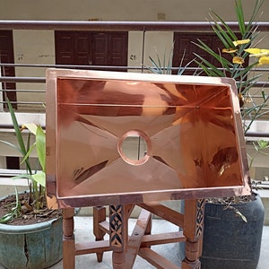 May include: A copper sink with a round drain hole sits on a wooden stand with intricate carvings. The sink is polished and reflects the surrounding environment.