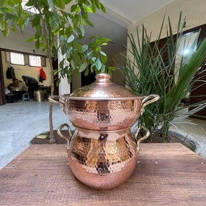 Pode incluir: Uma panela de cobre com acabamento martelado e duas alças. A panela tem uma tampa com uma pega no topo. A panela está sobre uma superfície de madeira.