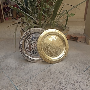 Puede incluir: Dos bandejas redondas de metal con diseños intrincados. Una bandeja es dorada con un patrón floral, y la otra es plateada con un patrón geométrico.