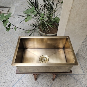 May include: A rectangular kitchen sink made of brushed brass with a metallic gold finish. The sink features a deep basin with angled bottom panels leading to a central circular drain assembly. The surface has a smooth, matte texture.