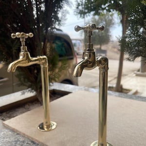 Könnte beinhalten: Zwei goldfarbene Wasserhähne im Wasserhahn-Stil mit einem kreuzförmigen Griff an jedem Auslauf. Die Ausläufe sind an hohen, zylindrischen Rohren befestigt, die ebenfalls goldfarben sind. Die Rohre sind auf einer hellbraunen Oberfläche montiert.