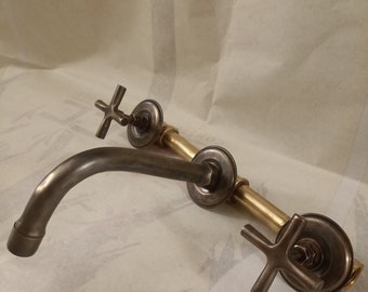 May include: A vintage-style faucet with a curved spout and cross-shaped handles. The faucet features a bronze finish on the spout and handles, with brass-colored connecting pipes. The design evokes a classic, antique aesthetic.