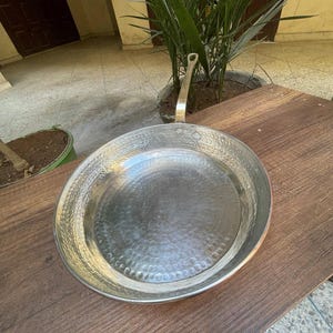 May include: A silver hammered copper pan with a long handle. The pan is sitting on a wooden surface.