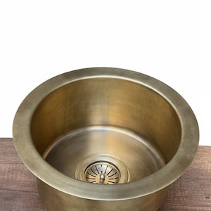 May include: A circular, antique brass kitchen sink with a drain. The sink has a smooth, slightly textured surface and a wide rim. The drain features a decorative, slotted design. The sink is set against a wooden surface.