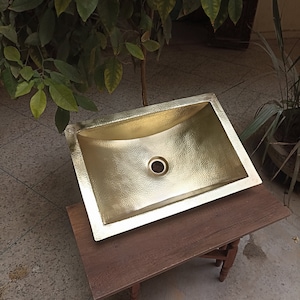 Puede incluir: Un lavabo rectangular dorado martillado con un desagüe central. El lavabo está colocado sobre una pequeña mesa de madera oscura. La superficie del lavabo tiene un aspecto texturizado. El fondo incluye follaje verde y una superficie de hormigón.