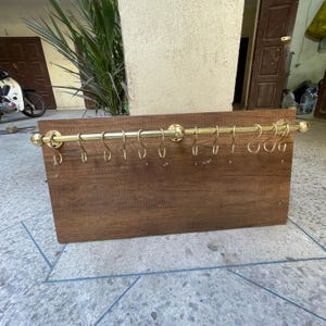 Puede incluir: Un estante de pared de madera con una barra y ganchos de latón. El estante tiene un acabado de madera marrón oscuro y está diseñado para sostener utensilios de cocina u otros artículos.
