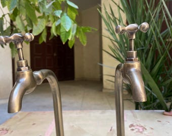 Puede incluir: Dos grifos de latón de estilo antiguo con manijas en forma de cruz, montados sobre una superficie de madera. Los grifos tienen un caño alto y curvo y una base con un diseño decorativo.