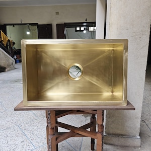 Peut inclure: Un évier de cuisine rectangulaire avec une finition dorée brossée et une ouverture de vidange circulaire centrale. L'évier présente des bords droits et nets ainsi qu'une surface métallique lisse qui reflète la lumière, révélant un bassin profond.