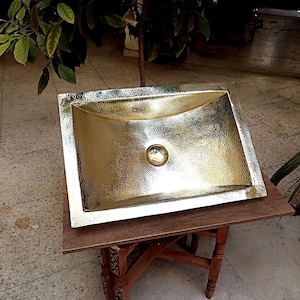 Könnte beinhalten: Ein gehämmertes, goldfarbenes rechteckiges Waschbecken mit abgerundetem Innenraum. Das Waschbecken steht auf einem kleinen, dunklen Holztisch mit dekorativen Beinen. Das Waschbecken hat eine strukturierte Oberfläche und einen zentralen Abfluss.