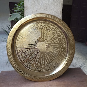 以下が含まれることがあります： 複雑な彫刻が施された、大きな丸い金色の金属製のサービングトレイ。トレイは縁が高く、装飾的な縁取りが施されています。