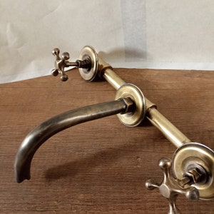 May include: A brass faucet with two cross-shaped handles and a curved spout. The faucet is mounted on a wall and has a pipe-like design.