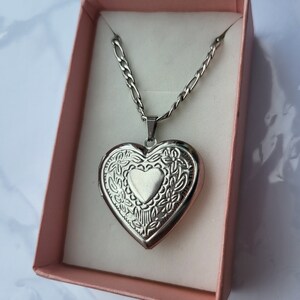 May include: A silver heart-shaped locket necklace with a delicate chain. The locket features an intricate floral design and a smaller heart in the centre. The necklace is displayed in a pink box.