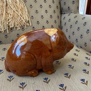May include: A brown ceramic pig-shaped bank with a glossy finish. The bank has a slot on top for inserting coins and is sitting on a patterned chair. The pig has a rounded body and small legs.
