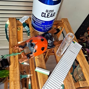 Op de afbeelding: Een houten decoratieve ladder met een lieveheersbeestje beeldje, een transparante spuitbus en een genummerd papier. De spuitbus leest "GLOSS CLEAR". Het lieveheersbeestje is rood en zwart.