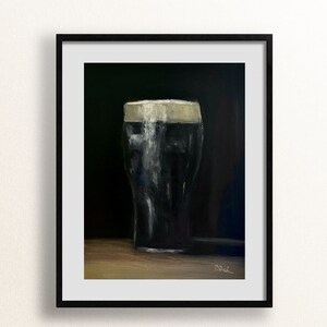 May include: A framed art print of a pint of dark stout beer with a creamy white head. The artwork is a painting with a dark background and a wooden surface. The glass is filled with a dark liquid, and the foam is thick and textured.