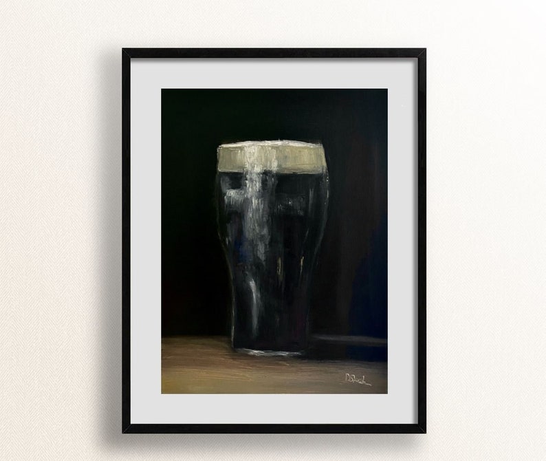 May include: A framed art print of a pint of dark stout beer with a creamy white head. The artwork is a painting with a dark background and a wooden surface. The glass is filled with a dark liquid, and the foam is thick and textured.