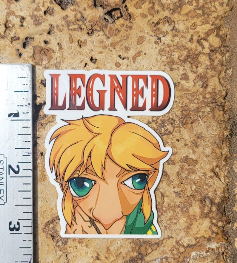 Peut inclure: Un autocollant avec une illustration de dessin anim&eacute; d'un personnage aux cheveux blonds, aux yeux verts et au nez pro&eacute;minent. Le personnage porte une chemise verte. Le mot "LEGNED" est imprim&eacute; en rouge au-dessus du personnage.