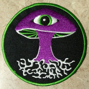 May include: A round embroidered patch with a green border. The patch features a purple mushroom with a green eye on top. The mushroom has white roots.