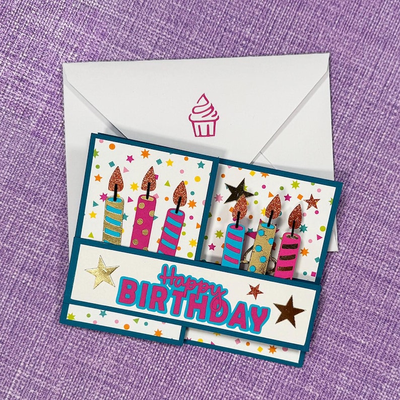 May include: A handmade birthday card with a white envelope featuring a pink cupcake stamp. The card has a teal border and displays the words "Happy Birthday" with colorful candles and star accents.