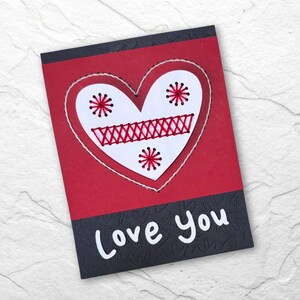 Pode incluir: Um cartão de Dia dos Namorados feito à mão com um coração branco costurado com linha vermelha sobre um fundo vermelho. O coração é decorado com explosões de estrelas vermelhas. As palavras "Love You" estão escritas em branco em uma borda preta estampada.