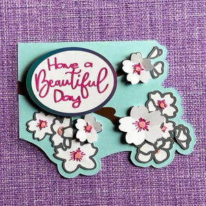 May include: A handmade greeting card with a light blue background and a brown branch design. The card features white paper flowers with pink centers and the text "Have a Beautiful Day" in pink script.