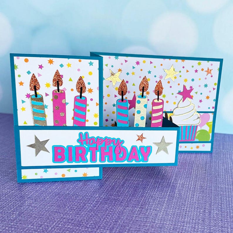 May include: A handmade birthday card with a pop-up design. The card features colorful candles with glitter flames, stars, and a cupcake. The words "Happy Birthday" are prominently displayed in pink and blue.
