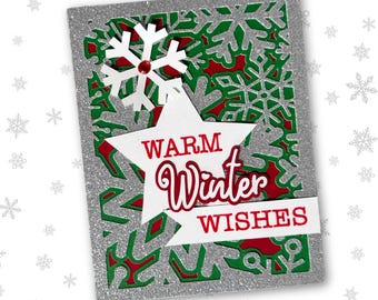 Puede incluir: Una tarjeta navideña plateada brillante con un diseño en capas. La tarjeta presenta una estrella blanca con las palabras "WARM Winter WISHES" en rojo. Un copo de nieve blanco con una gema roja está en la parte superior. El fondo tiene diseños recortados verdes y rojos.