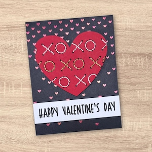 May include: A Valentine's Day card with a red heart stitched with white and gold thread, featuring "XOXO" designs. The card has a dark grey background with small pink hearts and a white banner that reads "HAPPY VALENTINE'S DAY."
