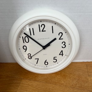 May include: White round wall clock with black numerals and hands. The clock face has the word "INGRAHAM" printed in the center. The clock is on a wooden surface.