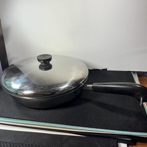 May include: Stainless steel frying pan with a lid and a black handle. The pan is round and has a shiny, reflective surface. The lid has a black knob. The handle is attached to the side of the pan.
