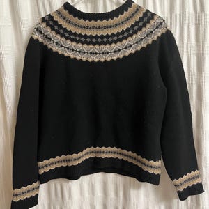 May include: Black sweater with a patterned yoke and hem. The yoke features a repeating design in tan, gray, and off-white. The sweater is hanging on a white hanger against a white curtain background.