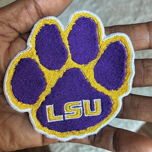 Peut inclure: &Eacute;cusson brod&eacute; en forme de patte, violet avec un contour dor&eacute;. Les lettres "LSU" sont brod&eacute;es en or &agrave; l'int&eacute;rieur de la patte. Id&eacute;al pour les v&ecirc;tements ou accessoires.
