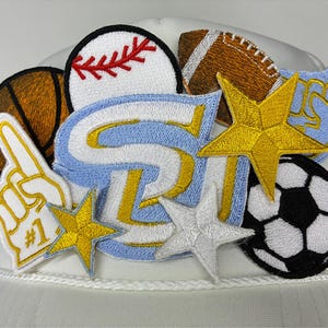 Op de afbeelding: Witte baseballcap met geborduurde patches van sportuitrusting en sterren. Patches zijn onder meer een honkbal, een American football, een basketbal en een voetbal. De patches zijn in verschillende kleuren.