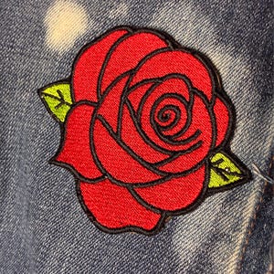 May include: An embroidered patch featuring a vibrant red rose with black outlines and two bright green leaves. The rose is stitched with dense thread, showing detailed petal layers and a central spiral, attached to a blue denim fabric background.