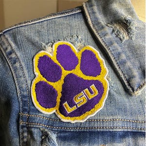 Peut inclure: &Eacute;cusson brod&eacute; en forme de patte de couleur violette et dor&eacute;e avec les lettres "LSU" brod&eacute;es en or. L'&eacute;cusson est cousu sur une veste en jean bleu clair.
