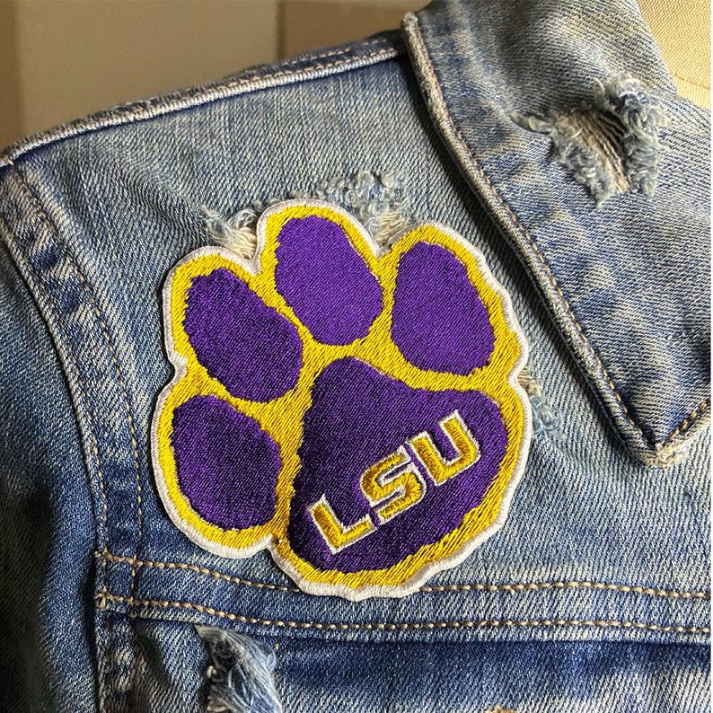 Peut inclure: &Eacute;cusson brod&eacute; en forme de patte de couleur violette et dor&eacute;e avec les lettres "LSU" brod&eacute;es en or. L'&eacute;cusson est cousu sur une veste en jean bleu clair.