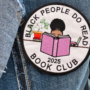 May include: Circular embroidered patch with the text "BLACK PEOPLE DO READ BOOK CLUB 2025". The design features a person reading a pink book, a potted plant, and other books. The patch is sewn onto a denim jacket.