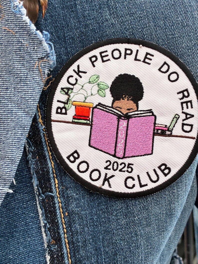 May include: Circular embroidered patch with the text "BLACK PEOPLE DO READ BOOK CLUB 2025". The design features a person reading a pink book, a potted plant, and other books. The patch is sewn onto a denim jacket.