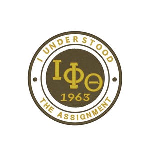 May include: A circular embroidered patch with the text "I UNDERSTOOD THE ASSIGNMENT" in gold lettering. The center features Greek letters and the year 1963, all in gold. The patch has a brown and white color scheme.