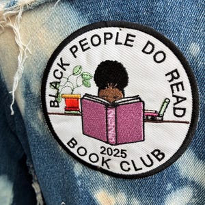 May include: A circular patch embroidered with the words "BLACK PEOPLE DO READ BOOK CLUB 2025". The patch shows a person reading a book, a plant, and other books. The patch is attached to a blue denim jacket.