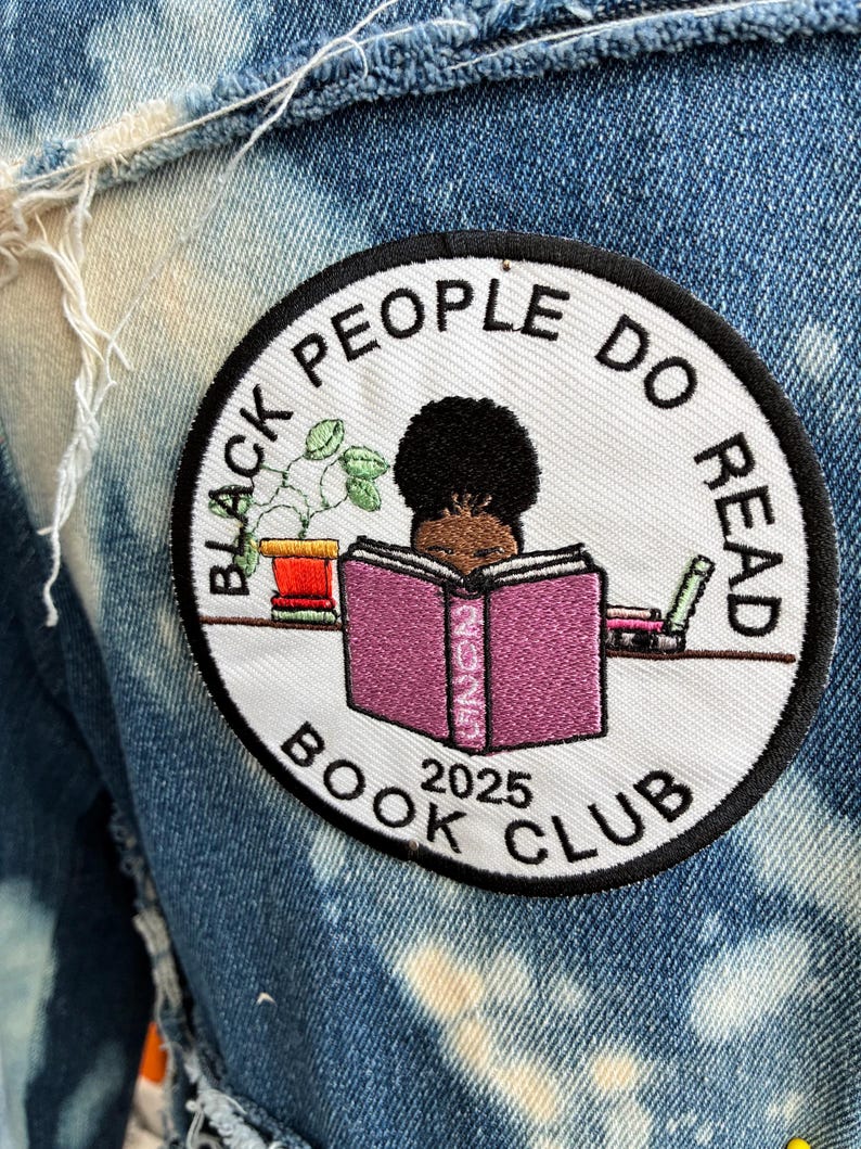 May include: A circular patch embroidered with the words "BLACK PEOPLE DO READ BOOK CLUB 2025". The patch shows a person reading a book, a plant, and other books. The patch is attached to a blue denim jacket.