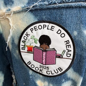May include: A denim jacket with a circular patch showing the text "BLACK PEOPLE DO READ BOOK CLUB 2025" around an illustration of a person reading. The jacket has a bleached, distressed design, with visible frayed edges.
